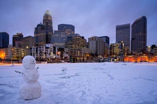 A snowman stands in a snowy landscape with a city skyline in the background during twilight.