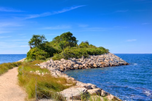 A scenic coastal view featuring a rocky shore, lush greenery, and a clear blue sky over calm waters.