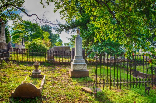 A serene cemetery scene with gravestones and a wrought iron fence, surrounded by lush green trees and grass.