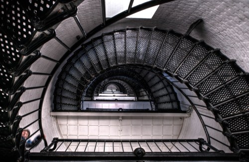 A spiral staircase viewed from above, showcasing intricate metalwork and a bright light source at the top.