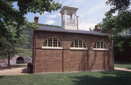 Historic brick building with arched windows and a tower, surrounded by trees and grassy area.