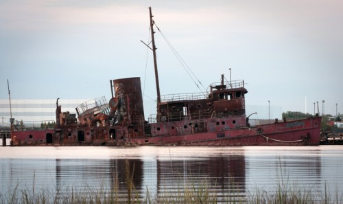 A rusted shipwreck sits partially submerged in calm waters, surrounded by tall grass and a cloudy sky.