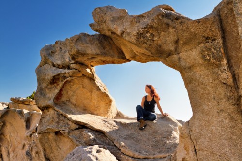 A woman sits on a rock formation with a natural arch, enjoying a sunny day against a clear blue sky.