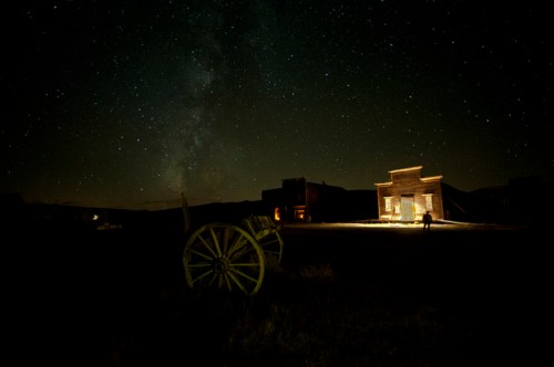 A starry night sky over an old western town with a wooden wagon in the foreground and a lit building in the background.