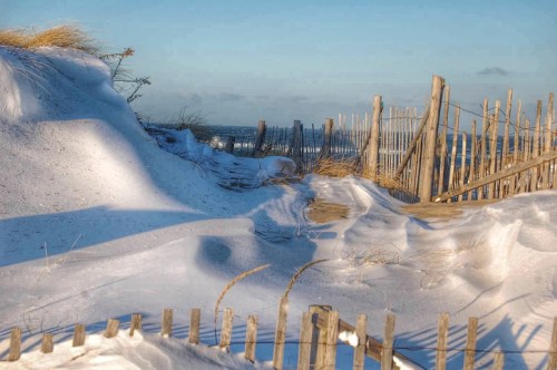 A sandy path leads through snow-covered dunes, with wooden fencing and a clear blue sky in the background.
