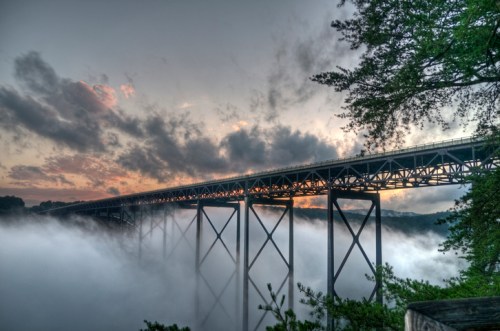 A bridge spans a foggy landscape at sunset, with clouds and trees framing the scene.