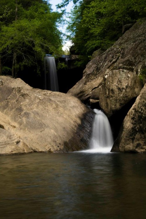 A serene landscape featuring two waterfalls cascading over rocky cliffs into a calm pool, surrounded by lush greenery.