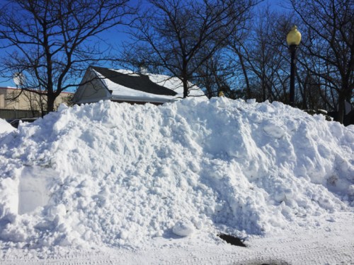 A large snow pile in a sunny area, with trees and a building partially visible in the background.