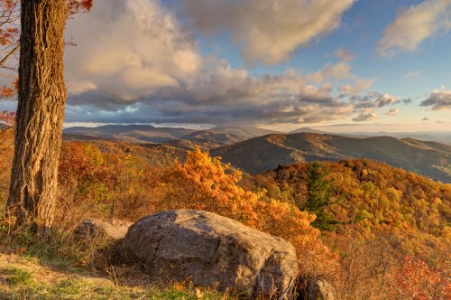 A scenic view of rolling mountains covered in autumn foliage under a partly cloudy sky. A large rock and tree are in the foreground.