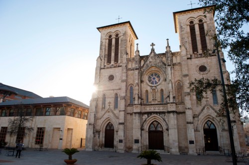 Historic church with twin towers and a rose window, bathed in sunlight, surrounded by a plaza and greenery.
