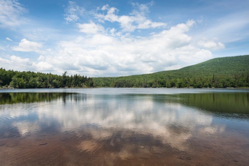 A serene lake reflects clouds and greenery, surrounded by mountains under a bright blue sky.
