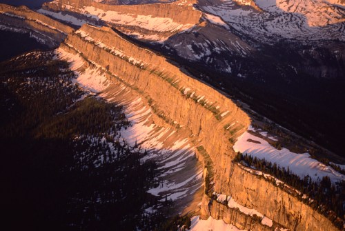 Aerial view of a rugged mountain ridge with snow-capped peaks and golden sunlight illuminating the landscape.