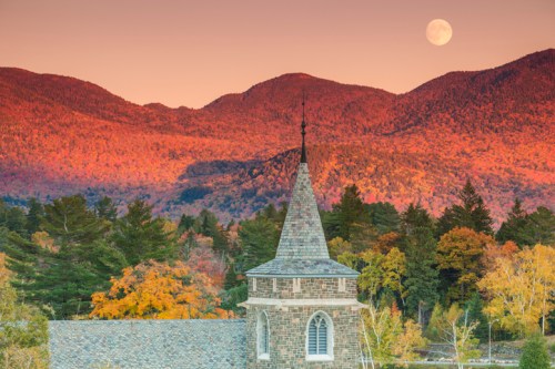 A stone church steeple in the foreground with vibrant autumn mountains and a full moon in the background.