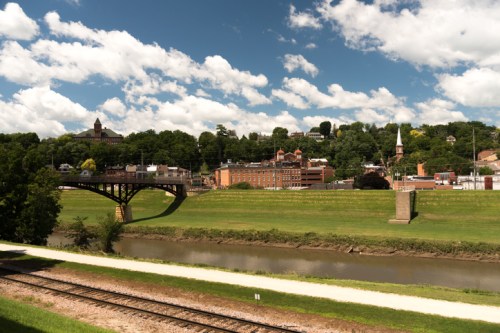 A scenic view of a town with a bridge over a river, surrounded by green hills and a blue sky with fluffy clouds.