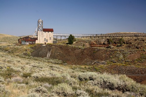 Abandoned mining structure on a hillside, with a wooden bridge and sparse vegetation under a clear blue sky.