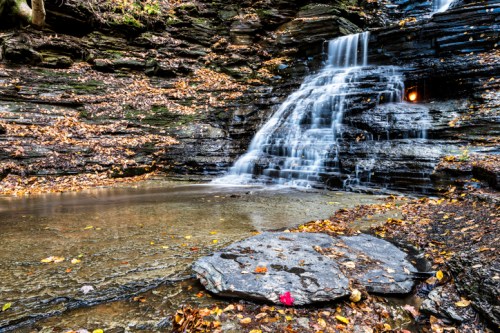 A serene waterfall cascades over rocky layers, surrounded by autumn leaves and a calm pool below.