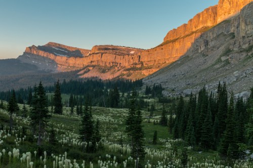 Sunlit mountain range at dawn, surrounded by lush green valleys and wildflowers under a clear blue sky.