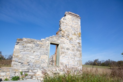 Ruins of a stone structure with a partially collapsed wall, set against a clear blue sky and grassy landscape.