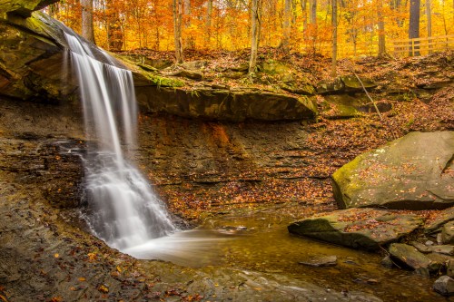 A serene waterfall cascades over rocks, surrounded by vibrant autumn foliage and a tranquil pool below.