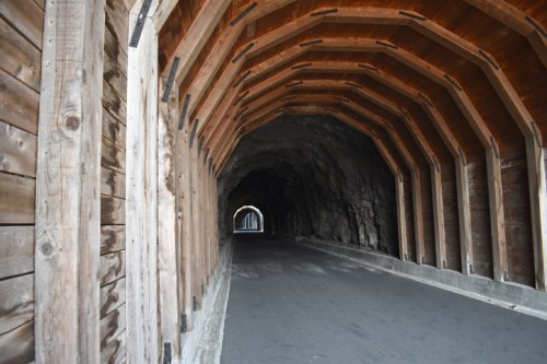 A wooden tunnel with a curved ceiling, leading into a rocky passageway, illuminated by natural light.