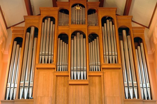 A large wooden pipe organ with multiple silver pipes, set against a warm-toned backdrop.