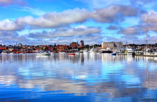 A serene harbor scene with boats reflecting on calm water, surrounded by buildings and a blue sky with clouds.