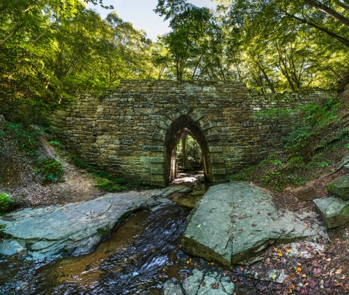 A stone arch bridge over a small stream, surrounded by lush green trees and rocky terrain.