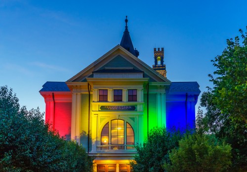 Historic building illuminated with rainbow colors at dusk, surrounded by trees and a clear blue sky.
