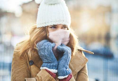 A woman in a winter coat, hat, and gloves, covering her face with a scarf, looks thoughtfully into the distance.