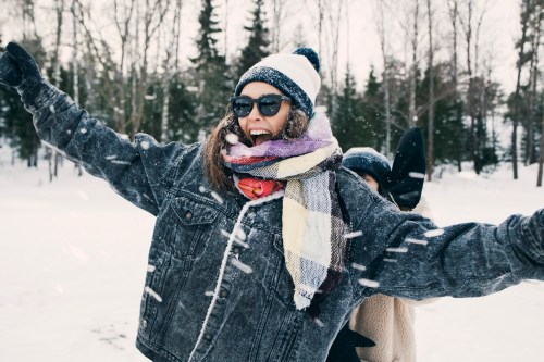 A joyful woman in sunglasses and a winter outfit smiles and spreads her arms in a snowy landscape.