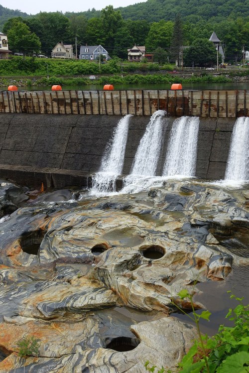 Water cascades over a dam, flowing onto rocky terrain with green hills and houses in the background.