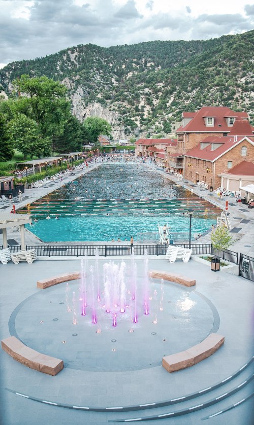 A scenic view of a large swimming pool with fountains, surrounded by mountains and historic buildings.