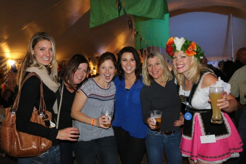 A group of six women smiling and posing together at a festive event, holding drinks under a decorated tent.