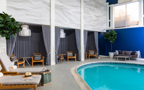 Indoor pool area with lounge chairs, curtains, and plants, featuring a blue wall and a serene atmosphere.