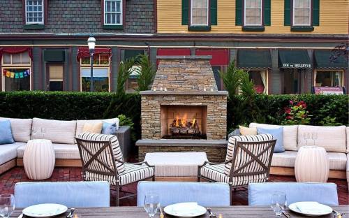 Cozy outdoor seating area with a stone fireplace, surrounded by greenery and colorful buildings in the background.