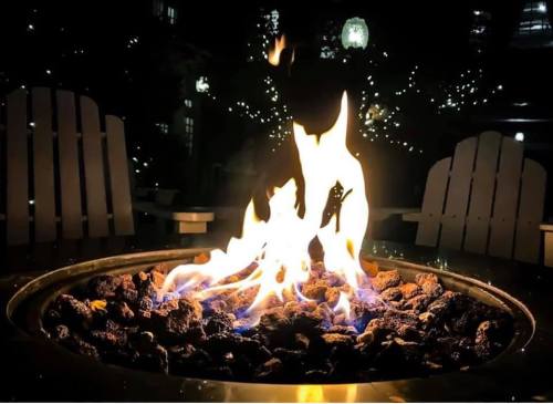 A close-up of a fire pit with flames dancing over rocks, surrounded by soft evening lights and wooden chairs.