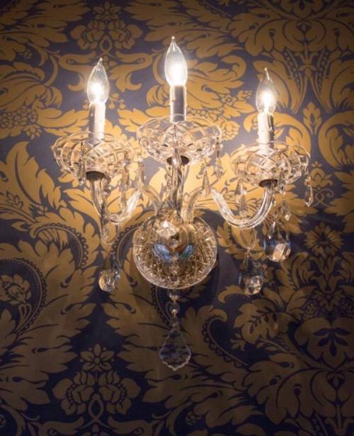 A crystal chandelier with three lights against a patterned gold wallpaper background.