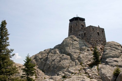 A stone tower atop a rocky hill, surrounded by trees and a clear sky. Steps lead up to the structure.