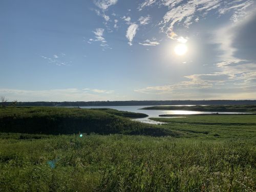 A serene landscape featuring a river, lush green fields, and a bright sun in a partly cloudy sky.