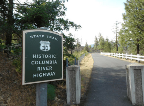 Sign for the Historic Columbia River Highway on a paved trail surrounded by trees and greenery.