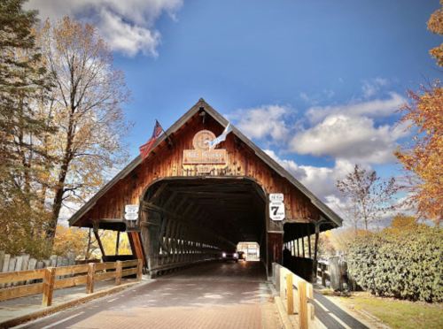A scenic view of a wooden covered bridge with autumn foliage and a cloudy sky in the background.