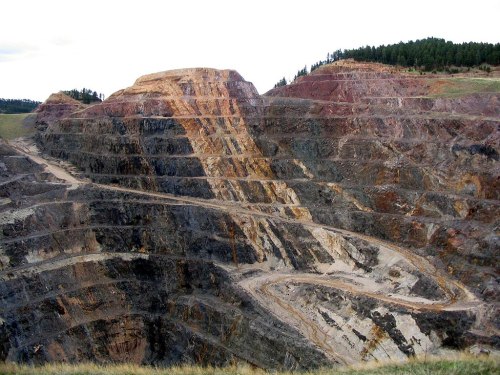 A large, multi-colored open-pit mine with steep, layered walls and a winding road leading down the slope.