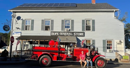 A red vintage fire truck parked in front of the Hope General Store, a blue building with solar panels on the roof.