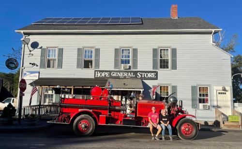 A vintage red fire truck parked in front of the Hope General Store, with two people sitting on its bumper.
