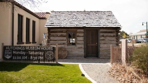 A small log cabin with a sign displaying museum hours, surrounded by grass and a pathway.