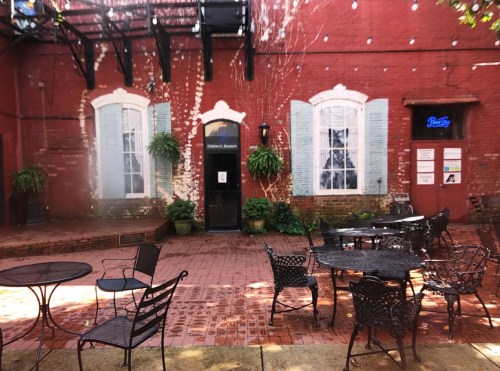 A quaint outdoor courtyard with metal tables and chairs, surrounded by red brick walls and green plants.