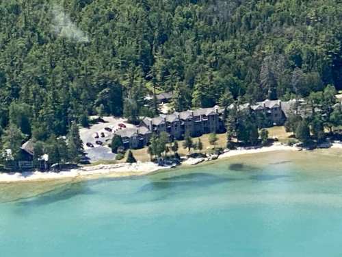 Aerial view of a lakeside property surrounded by trees, with a sandy shore and clear blue water.