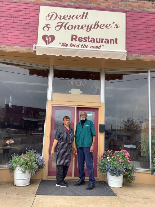 A woman in an apron and a man in a green shirt stand outside Drexell & Honeybee's Restaurant, which has a welcoming sign.