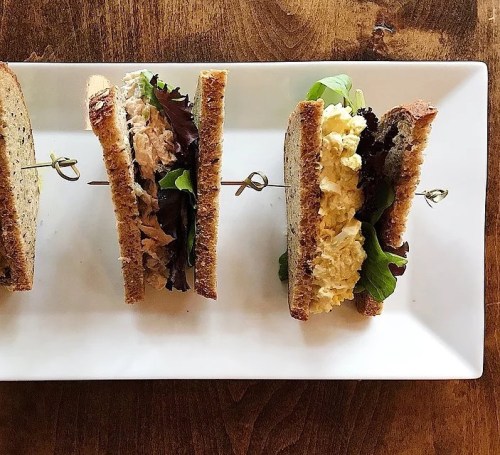 Three sandwiches on a white plate, each with different fillings, held together by skewers, on a wooden table.