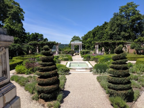 A serene garden with manicured hedges, a central fountain, and a clear blue sky in the background.